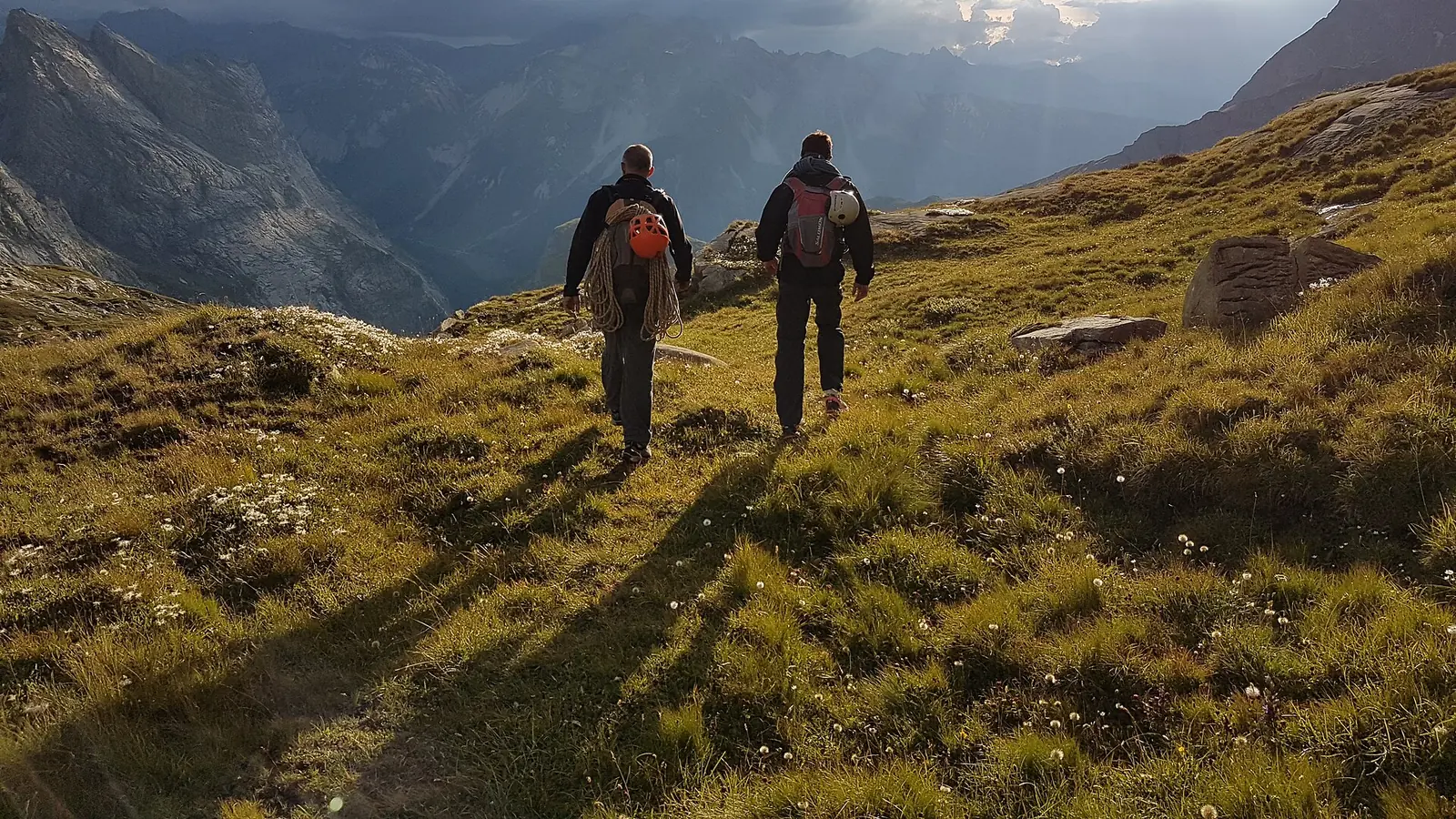 Two hikers walking down an alpine meadow into low evening sun, a deep valley and ridgelines behind them
